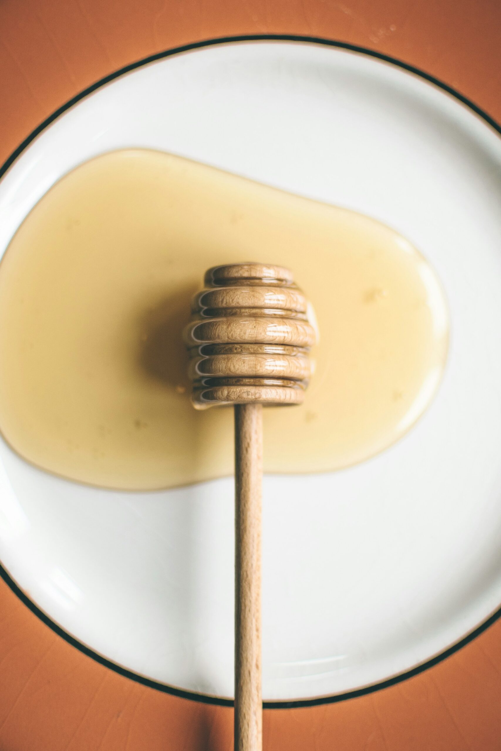 A wooden honey dipper resting in a pool of pale honey on a white plate with a dark rim, on an orange table