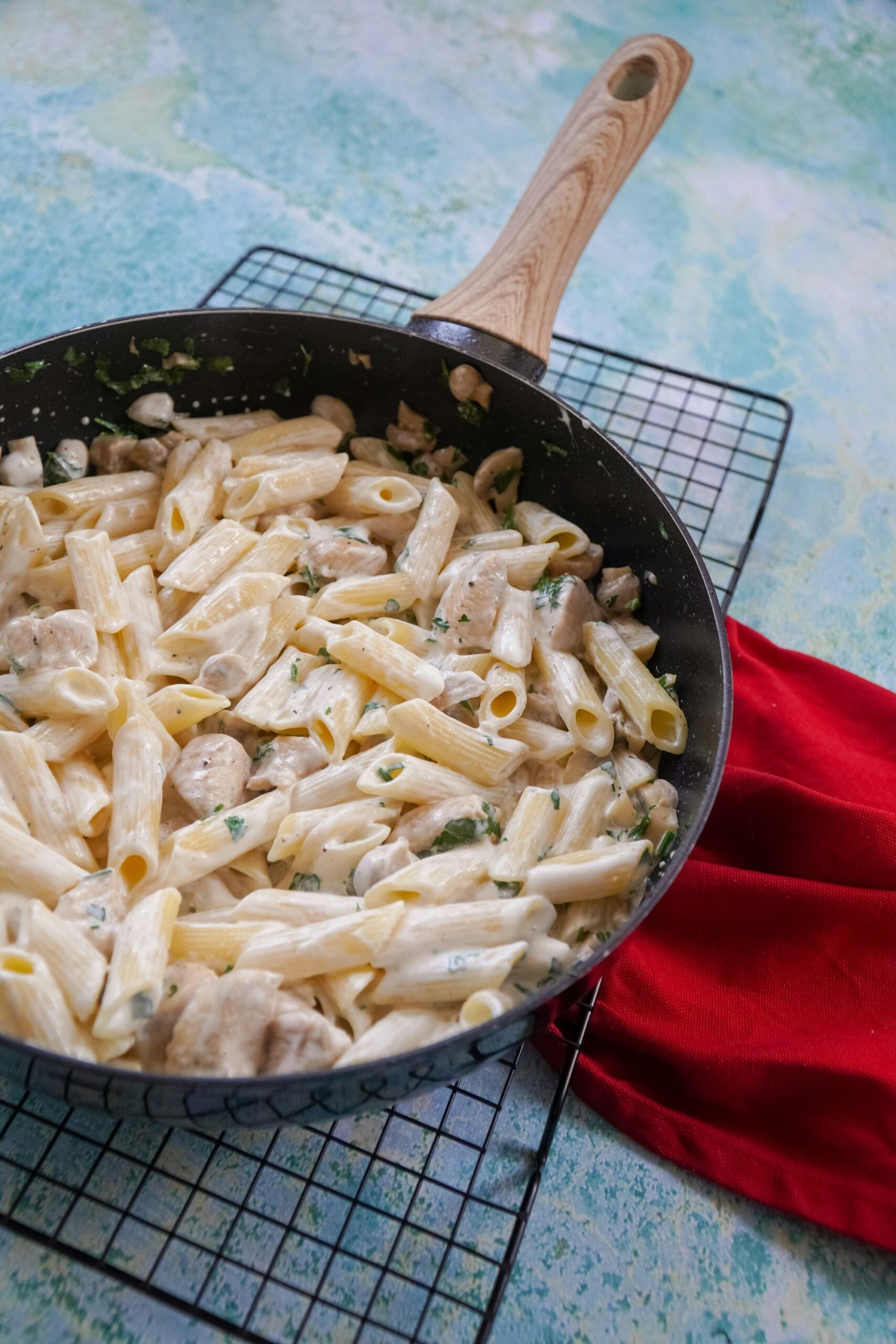 A black frying pan of penne in a creamy white sauce with pieces of chicken, mushrooms and chopped parsley, on a wire rack beside a red cloth
